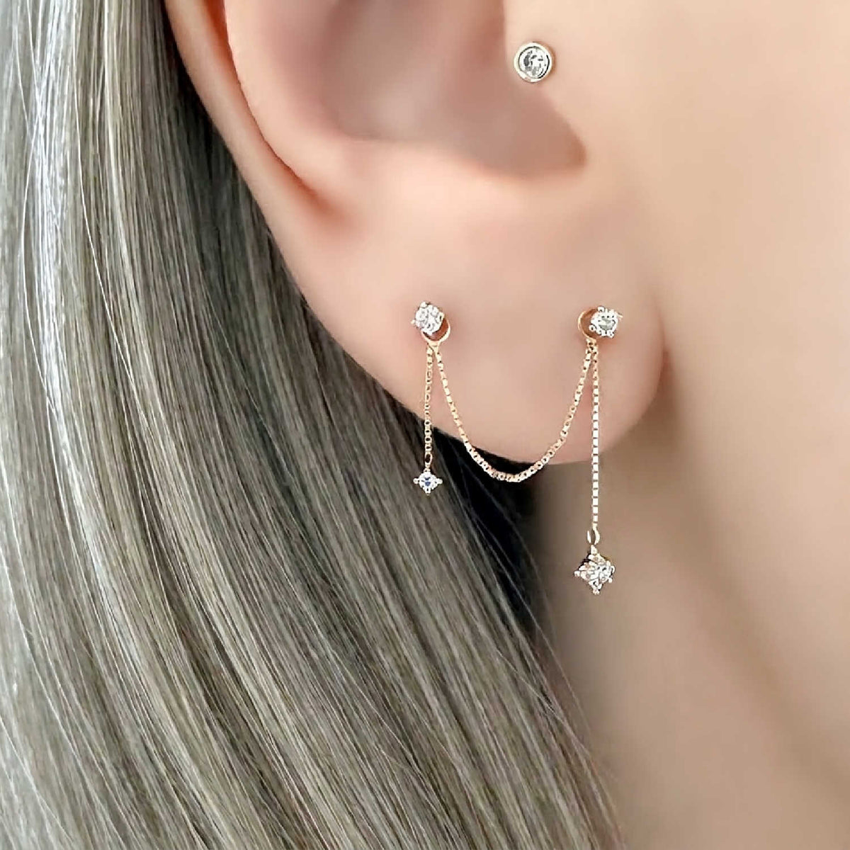 A close-up of a woman's ear wearing a double piercing dangling chain earring charm. The earrings are 14k solid gold with small diamond gemstones and thin chains