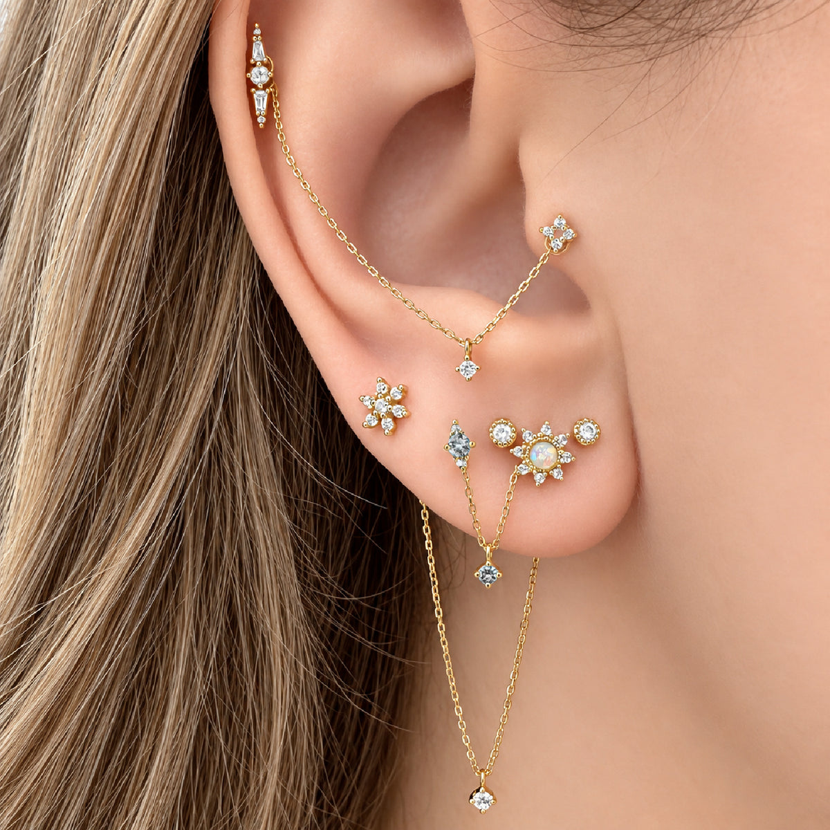 A close-up of a model's ear wearing a gold earring stack featuring blue topaz flat back studs and double piercing charms.