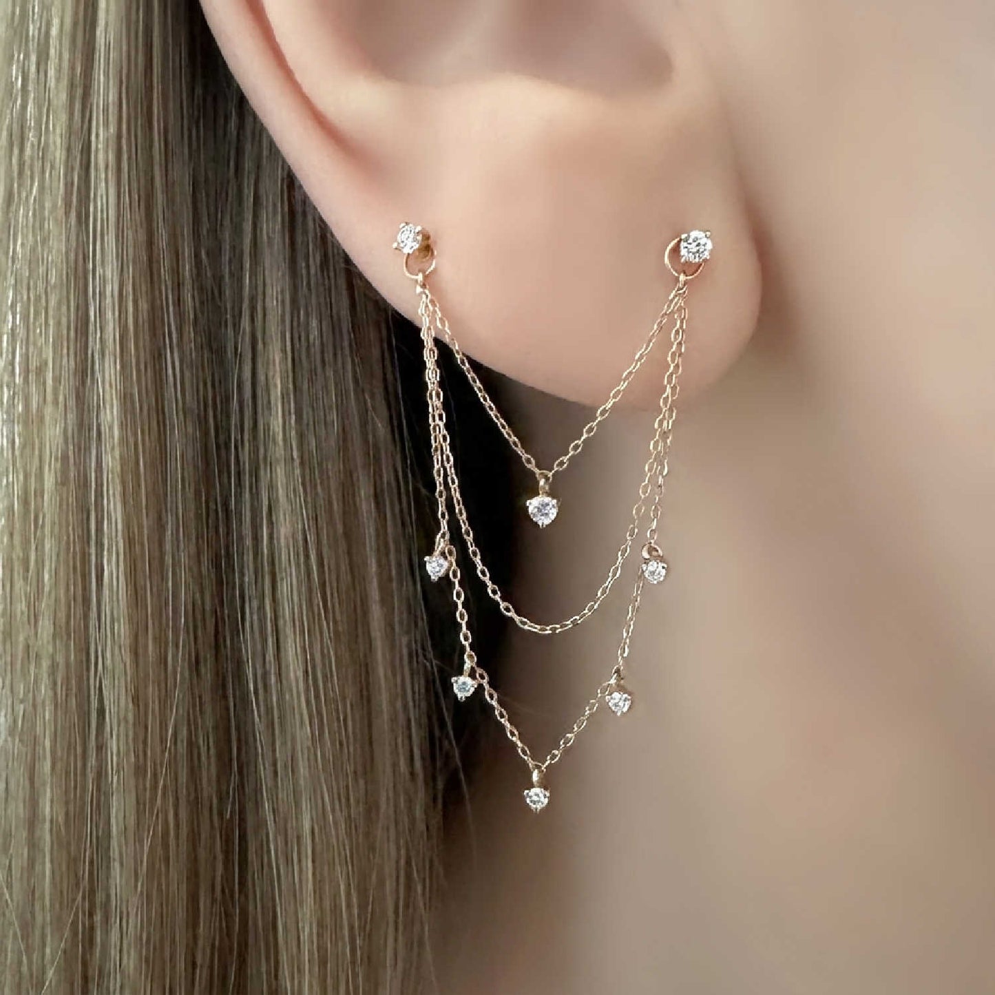 A close-up of a woman's ear wearing a double piercing dangling chain earring charm. The earrings are 14k solid gold with six diamond gemstones and 3 thin chains