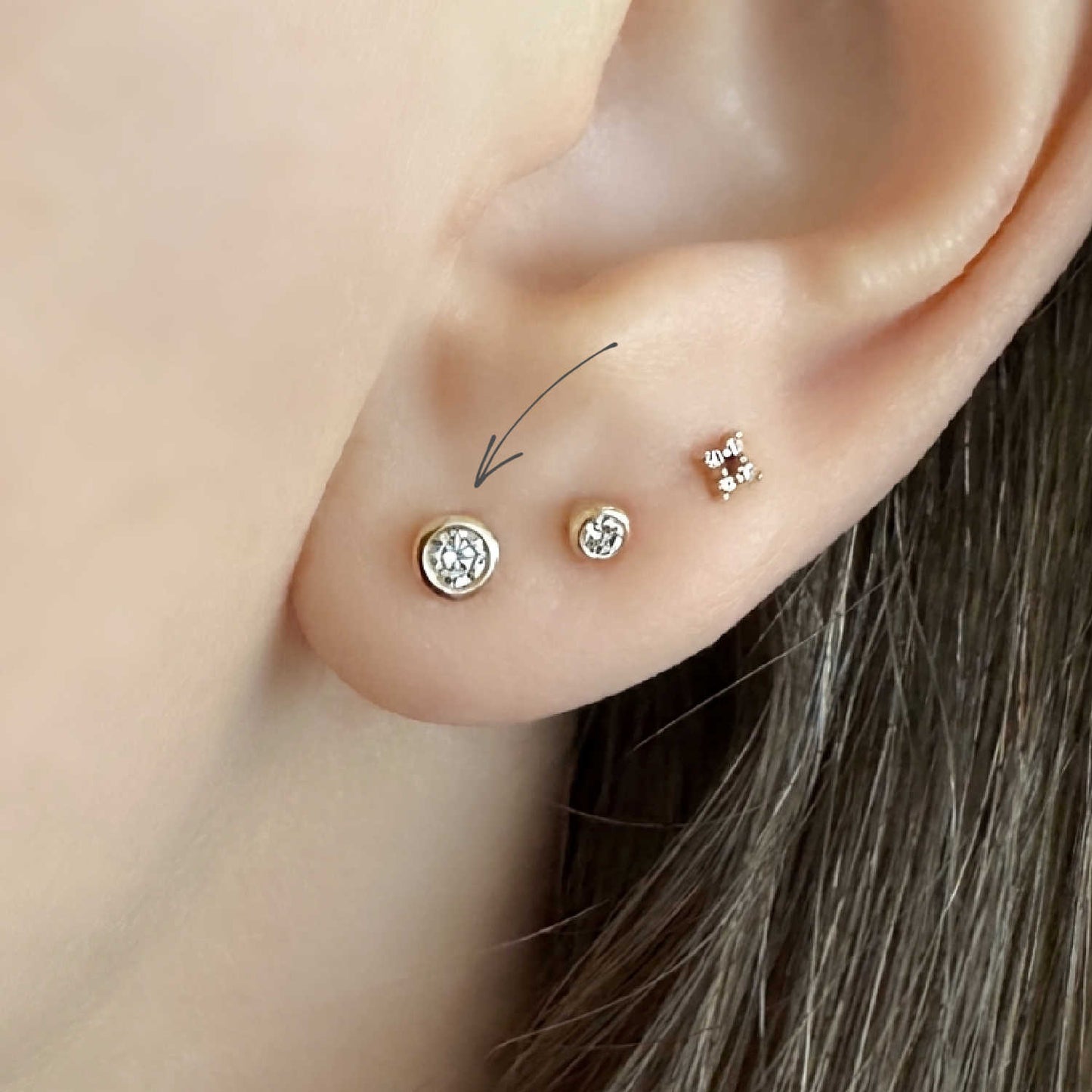 Close-up of a woman's ear showing three diamond stud earrings set in 14k solid gold bezels. A thin arrow points to the larger stud.