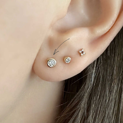 Close-up of a woman's ear showing three diamond stud earrings set in 14k solid gold bezels. A thin arrow points to the larger stud.