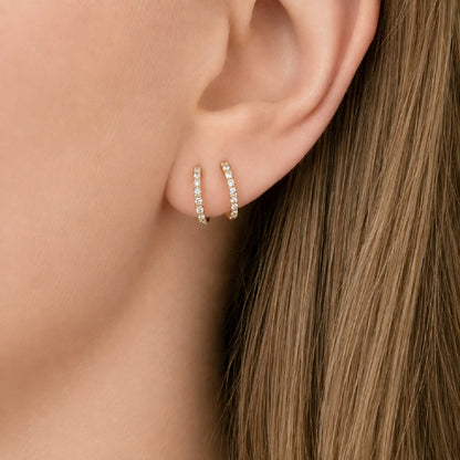 A close-up of a person's ear wearing a pair of thin 14k gold huggie hoop earrings set with diamonds, with long light brown hair visible.