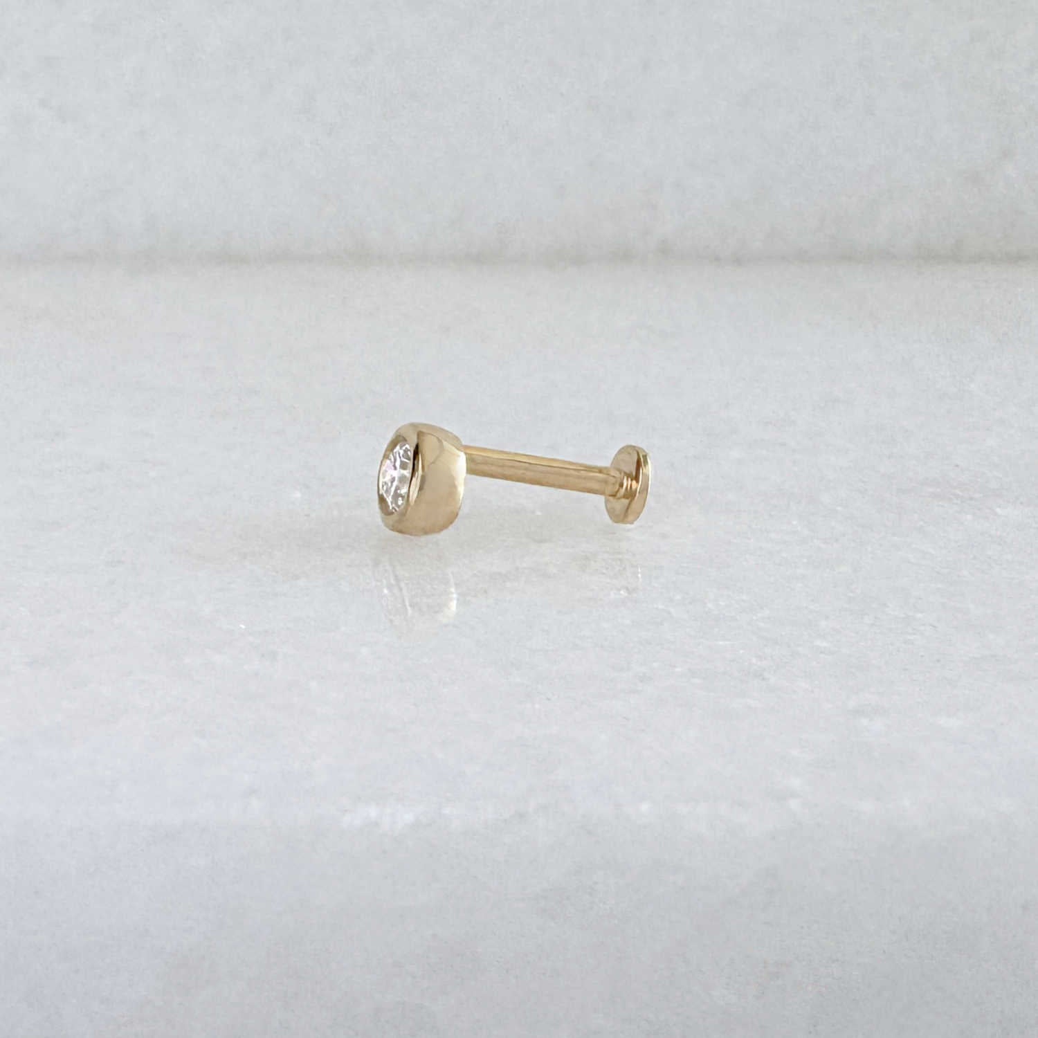Side view of a single 14k solid gold stud flat back earring with a bezel-set diamond gemstone, shown on a light gray background.