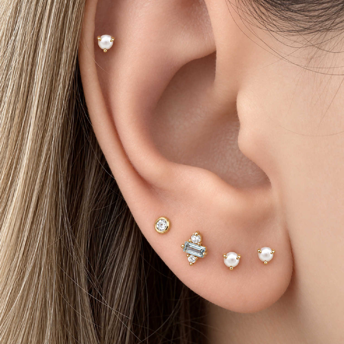 Close-up of a woman's ear showcasing a set of 14k gold ear piercings, including stud earrings with white pearls and a cluster with a blue topaz baguette gemstone.