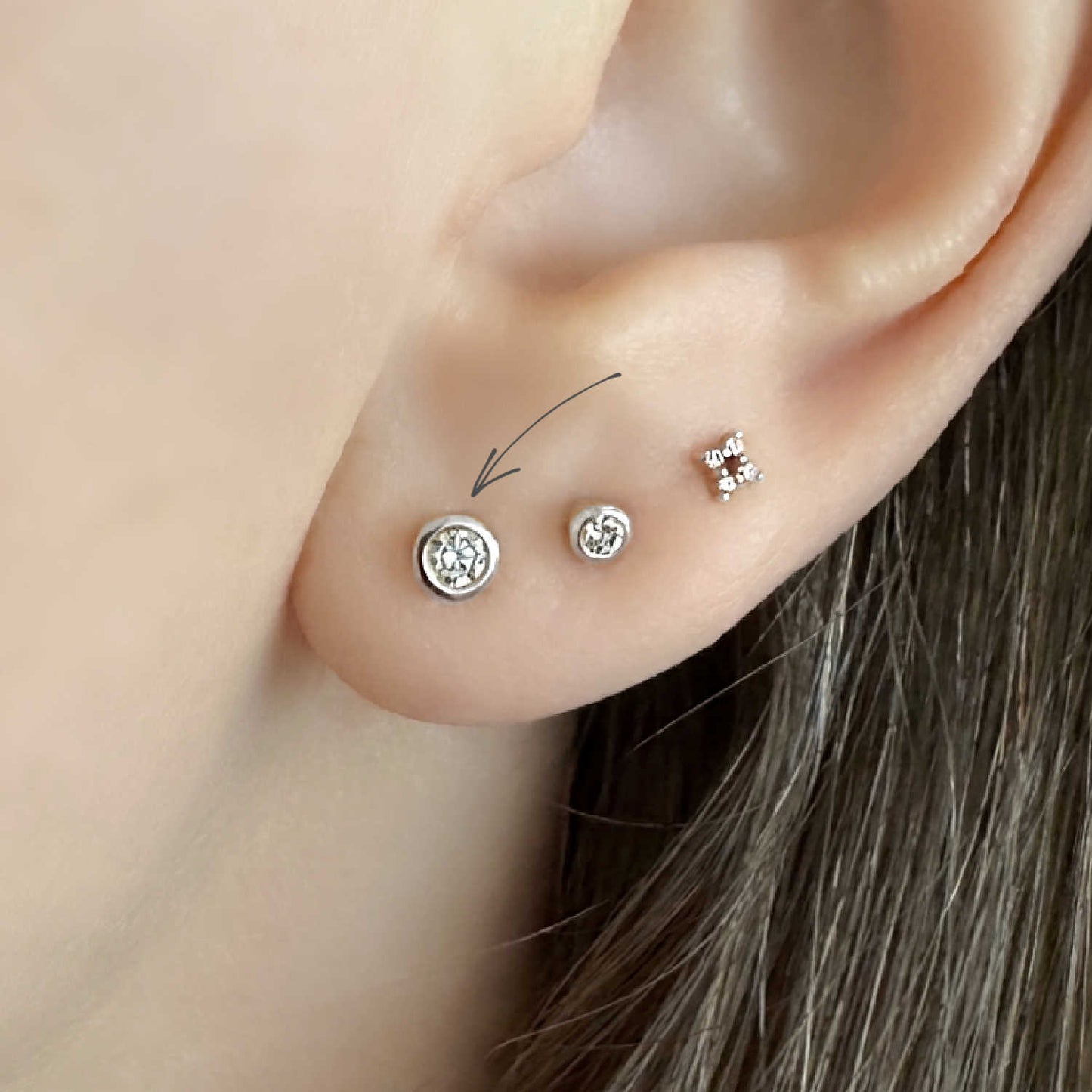 Close-up of a woman's ear showing three diamond stud earrings set in 14k solid white gold bezels. A thin arrow points to the larger stud.
