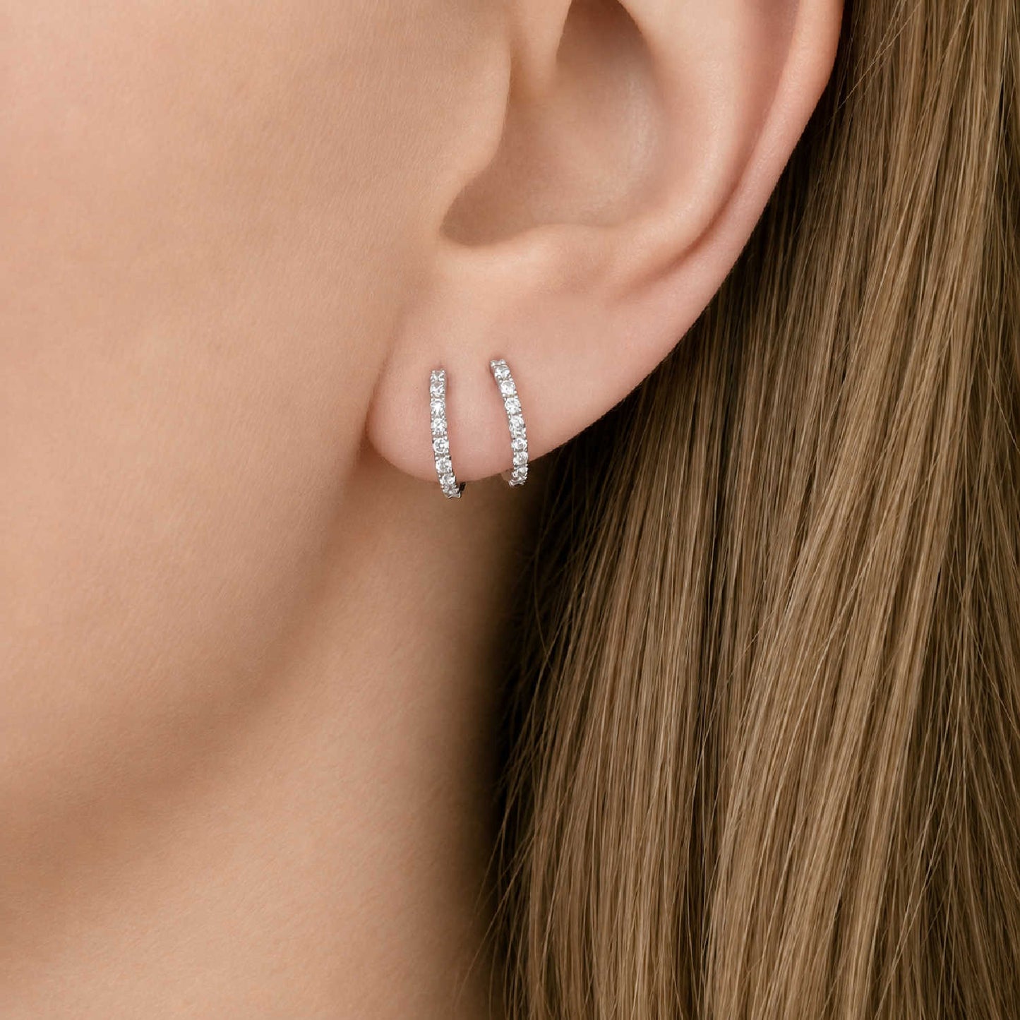 A close-up of a person's ear wearing a pair of thin 14k white gold huggie hoop earrings set with diamonds, with long light brown hair visible.