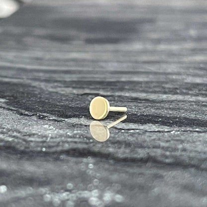 This image displays a single gold-colored flat back earring post, positioned on a textured grey surface. The earring post, simplistic and modern in design, features a smooth, round shape with a slight shine, highlighting its metallic finish.