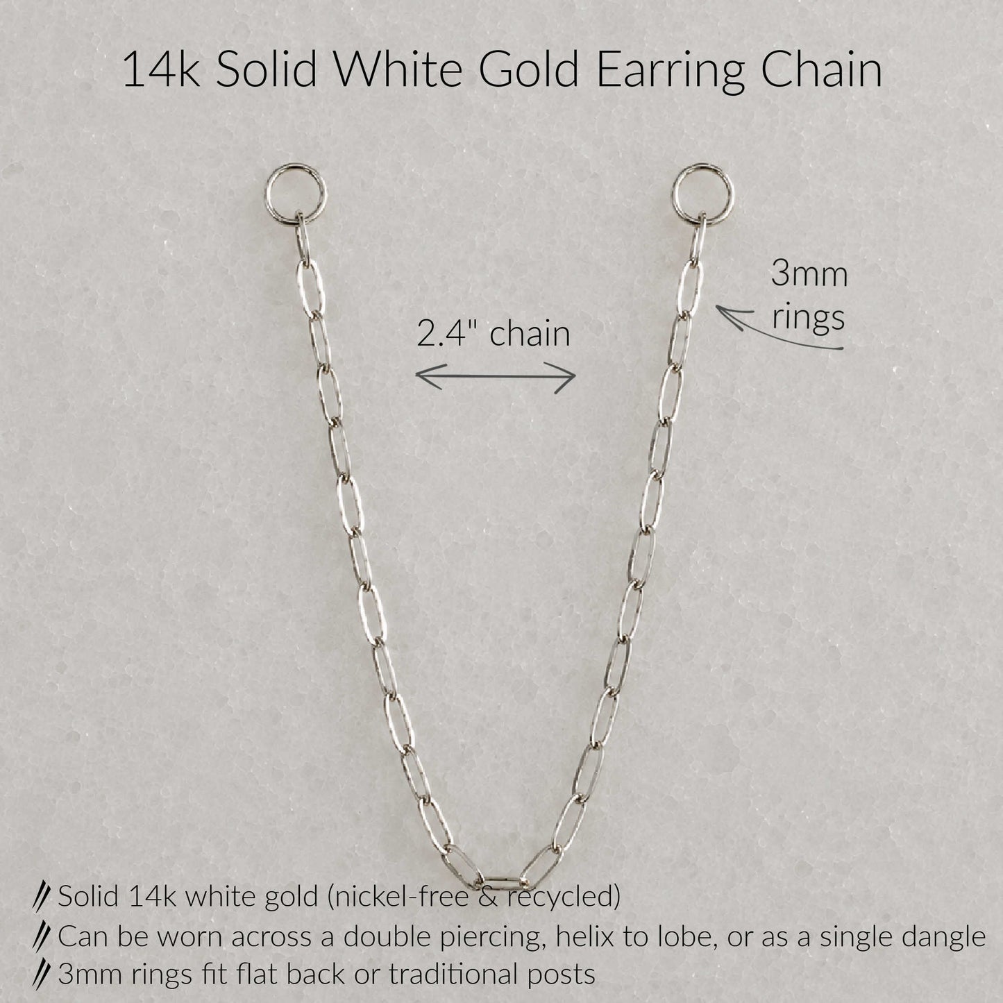 A 14k solid white gold earring chain shown on a light gray textured background with text annotations detailing its measurements and features: 2.4-inch chain length, 3mm rings, and product benefits including nickel-free recycled gold and multiple wear styles.