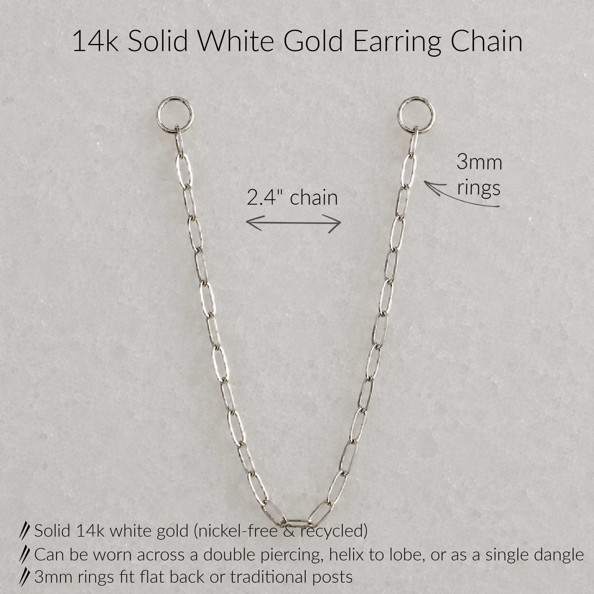 A 14k solid white gold earring chain shown on a light gray textured background with text annotations detailing its measurements and features: 2.4-inch chain length, 3mm rings, and product benefits including nickel-free recycled gold and multiple wear styles.