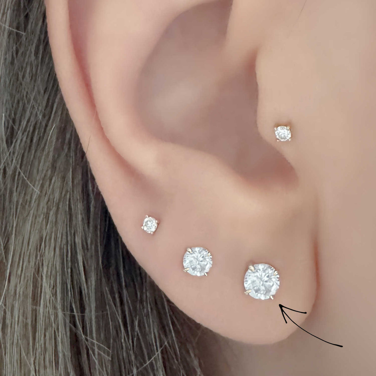 Close-up of an ear wearing four round diamond stud earrings in graduated sizes, with a black arrow pointing to the 1/3 carat stud.