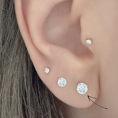 Close-up of an ear wearing four round diamond stud earrings in graduated sizes, with a black arrow pointing to the 1/3 carat stud.