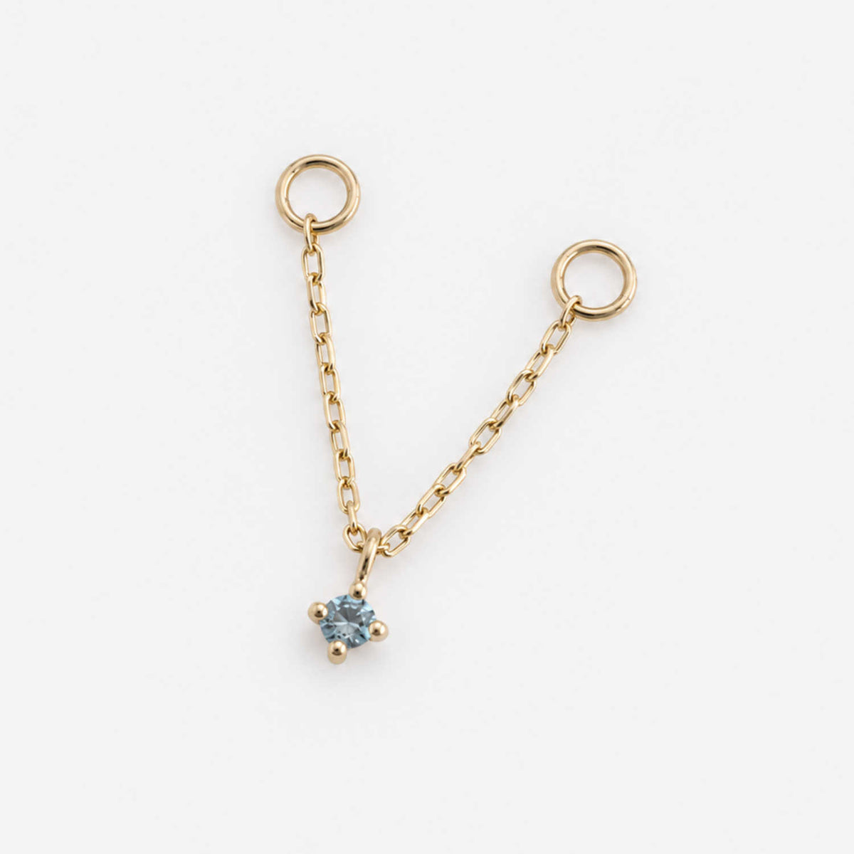 A delicate gold double piercing earring chain charm with two circular loops at the top and a small four-prong blue topaz, shown on a clean white background.