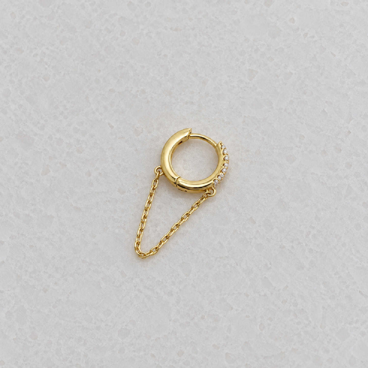 Side view of a small gold huggie hoop earring with a delicate chain drop and a row of tiny clear stones, photographed on a light textured surface.