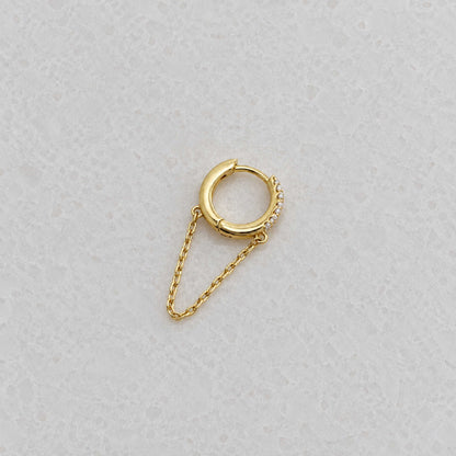 Side view of a small gold huggie hoop earring with a delicate chain drop and a row of tiny clear stones, photographed on a light textured surface.