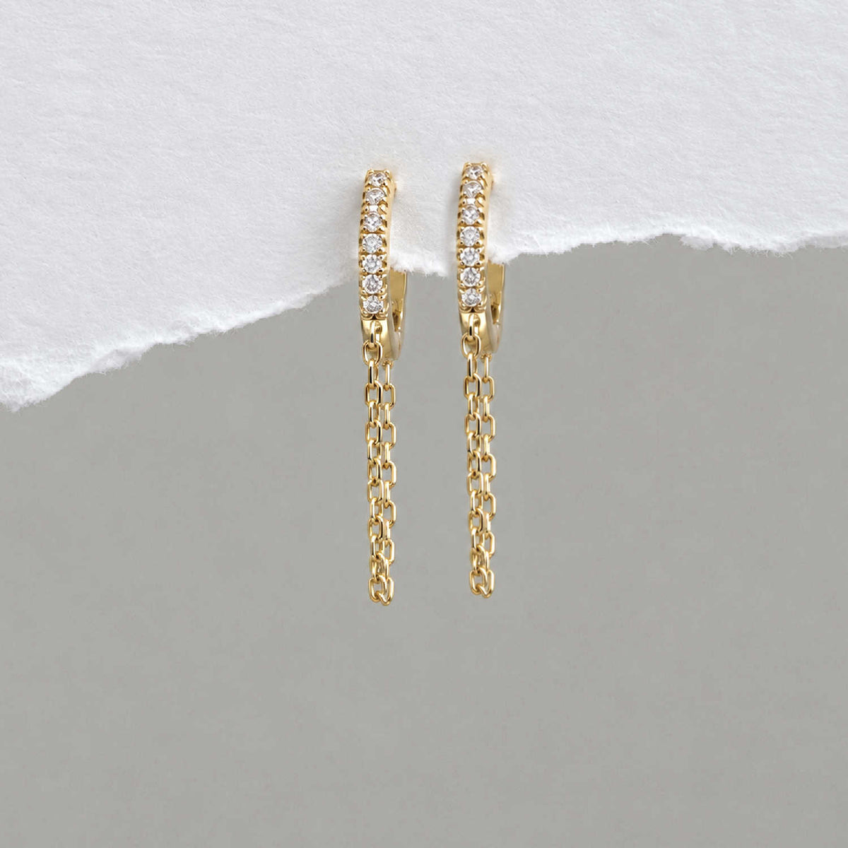 A pair of 14k gold plated huggie hoop earrings with a row of small clear cz stones and a dangling chain, displayed against a split white and gray background.