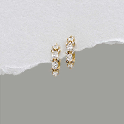A pair of 14k gold plated huggie hoop earrings with a vertical cluster of clear oval-cut and round stones, displayed on a textured white surface with a torn-paper effect over a gray background.