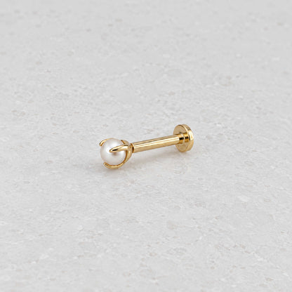 A single gold flat back stud piercing jewelry piece with a white pearl, resting on a light gray textured surface.