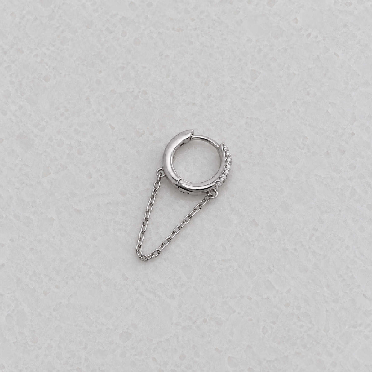 A silver huggie hoop earring with a small chain drop and a textured pavé accent on the side, displayed on a light gray textured surface.