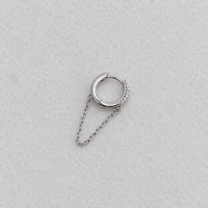 A silver huggie hoop earring with a small chain drop and a textured pavé accent on the side, displayed on a light gray textured surface.