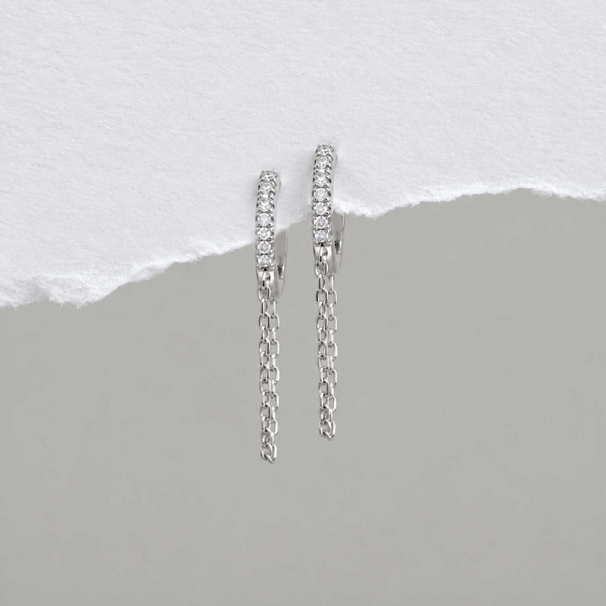 A pair of sterling silver huggie hoop earrings with a row of small clear cz stones and a dangling chain, displayed against a split white and gray background.