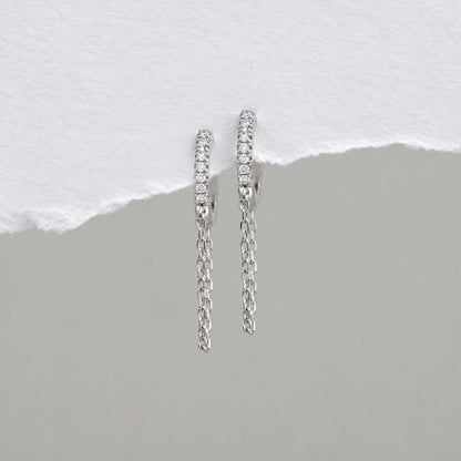 A pair of sterling silver huggie hoop earrings with a row of small clear cz stones and a dangling chain, displayed against a split white and gray background.