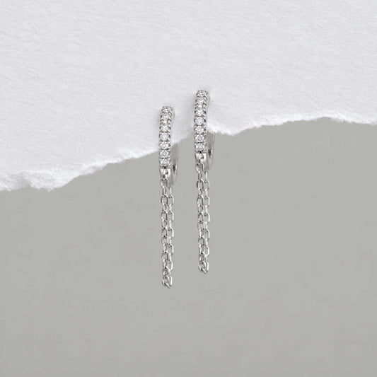 A pair of sterling silver huggie hoop earrings with a row of small clear cz stones and a dangling chain, displayed against a split white and gray background.