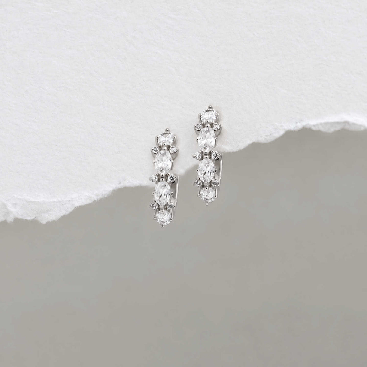 A pair of sterling silver huggie hoop earrings with a vertical cluster of clear oval-cut and round stones, displayed on a textured white surface with a torn-paper effect over a gray background.