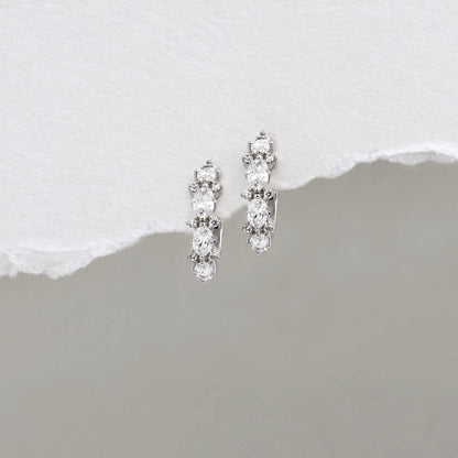 A pair of sterling silver huggie hoop earrings with a vertical cluster of clear oval-cut and round stones, displayed on a textured white surface with a torn-paper effect over a gray background.
