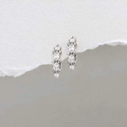 A pair of sterling silver huggie hoop earrings with a vertical cluster of clear oval-cut and round stones, displayed on a textured white surface with a torn-paper effect over a gray background.