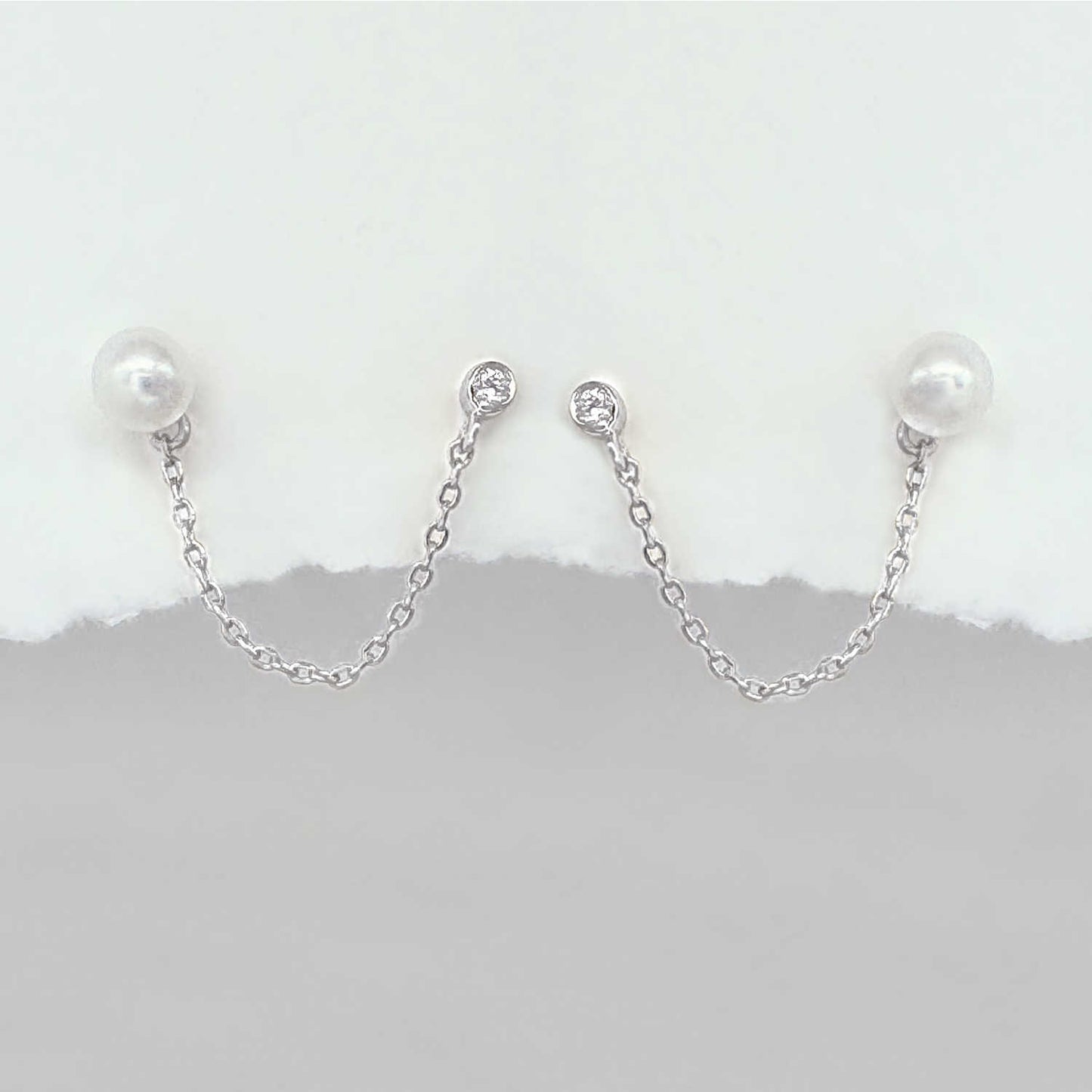 A pair of silver chain double piercing earrings with white pearl round studs and small clear stones at the top, displayed against a white and gray background.