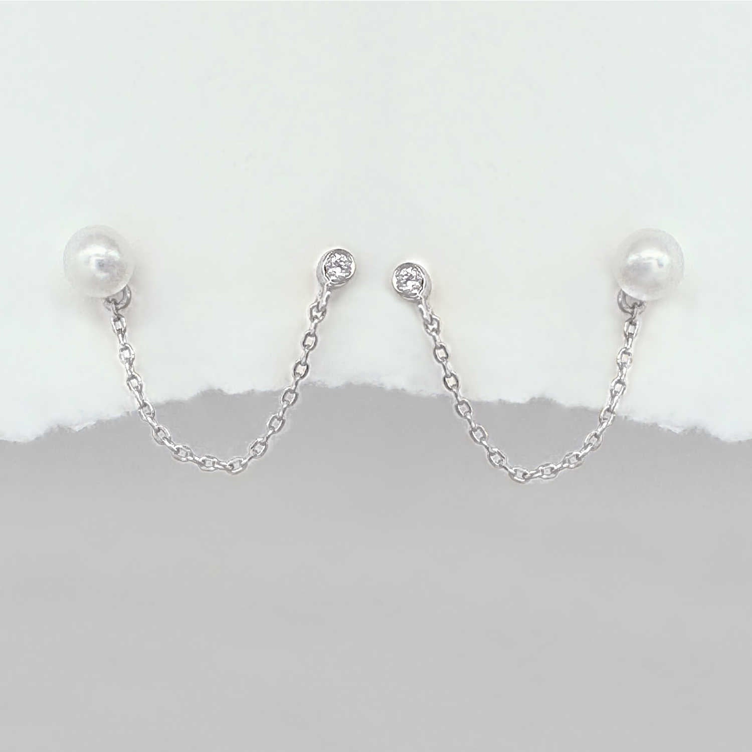 A pair of silver chain double piercing earrings with white pearl round studs and small clear stones at the top, displayed against a white and gray background.