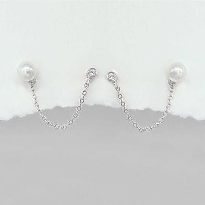 A pair of silver chain double piercing earrings with white pearl round studs and small clear stones at the top, displayed against a white and gray background.