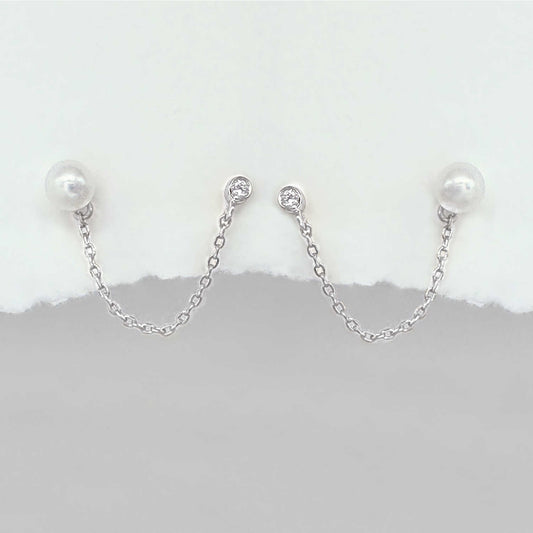 A pair of silver chain double piercing earrings with white pearl round studs and small clear stones at the top, displayed against a white and gray background.